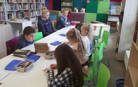 Dzieci i kobiety pracują przy stolikach.