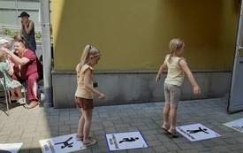 Dwie dziewczynki skaczą po kartkach, leżących na chodniku. Po lewej stronie grupa ludzi stoi wokół stolika.