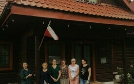 Grupa ludzi stoi przed drewnianym domem. W tle flaga Polski.