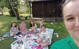 Dziewczyny siedzą na kocu, kobieta stoi, uśmiechają się do wspólnego zdjęcia.