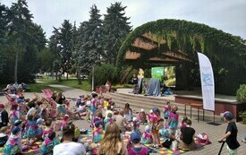 Grupa dzieci i dorosłych ogląda przedstawienie na plenerowej scenie w parku