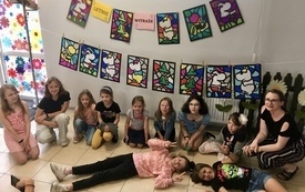 Grupa dzieci siedzi na podłodze, a nad nimi wisi galeria kolorowych witraży z postaciami Mumink&oacute;w