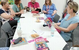 Grupa ludzi siedzi przy stole, na którym leżą książki, zakładki DKK i długopisy.