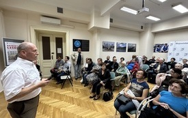 Sala konferencyjna. Z lewej strony mężczyzna w białej koszuli stoi twarzą do widowni. kt&oacute;ra z zainteresowaniem słucha. 