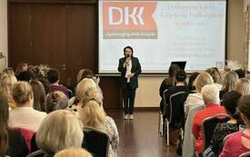Dyrektor biblioteki Bożena Janda trzyma w ręku mikrofon i mówi do zgromadzonych gości w przestrzennej sali. W tle ekran do wyświetlania prezentacji.