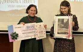 Dwie kobiety stoją obok siebie, jedna z nich trzyma bon podarunkowy, a druga zdjęcie oprawione w ramkę.