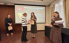 Kobieta przekazuje nagrodę na ręce drugiej kobiety. Obok stoi kobieta z mówi przez mikrofon. W tle czwarta kobieta trzyma torbę upominkową.