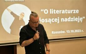 Radiowiec Łukasz Wojtusik trzyma w ręku mikrofon i mówi. W tle fragment slajdu prezentacji multimedialnej ze zdjęciem dziennikarza.