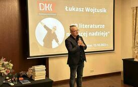 Dziennikarz Łukasz Wojtusik trzyma mikrofon i mówi. W tle slajd prezntacji multimedialnej z tytułem "O literaturze niosącej nadzieję".