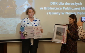 Dwie kobiety stoją, jedna trzyma bon podarunkowy, a druga prezentuje zdjęcie kobiet w ramce. 