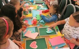 Dzieci siedzą przy długim stole i tworzą wycinanki z kolorowego papieru. 