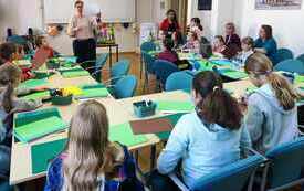 Grupa dzieci zgromadzona na warsztatach w bibliotece wraz z prowadzącą warsztaty kobietą w średnim wieku.