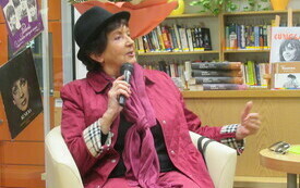 Kobieta w r&oacute;żowym stroju i czarnym kapeluszu, trzyma mikrofon w ręku.