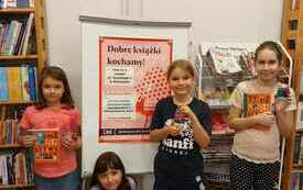 Cztery dziewczynki trzymają w rękach książki i słoiki. Za ich plecami plakat Dyskusyjnych Klubów Książki.