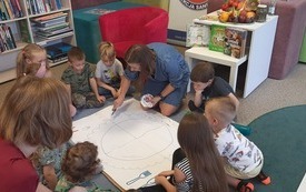 Dzieci i dorośli siedzą na dywanie wokół kartki papieru. Na papierze kobieta rysuje koło.