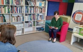 Bibliotekarka siedzi w fotelu i czyta książkę. Po lewej stronie regały z książkami.