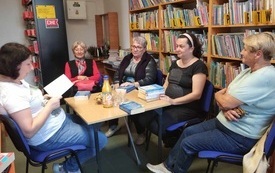 Pięć kobiet siedzi wokół stolika, jedna z nich trzyma książkę i czyta. Po prawej stronie regały z książkami.