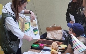 Dziewczyny stoją przy stole i patrzą w swoim kierunku.