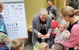 Kobieta pochyla się nad dziećmi stojącymi w kolejce, obok tablica.