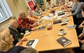 Wzdłuż stołu siedzą ludzie, na stole książki, talerze i filiżanki.
