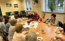 Grupa ludzi siedzi wokół stołu, na którym są filiżanki, talerzyki. Po prawej stronie okno.