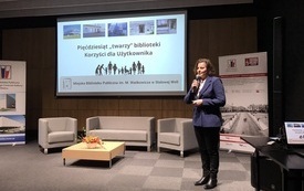 Kobieta w czarnych spodniach, białej koszuli i granatowej marynarce z mikrofonem w ręku. Na ekranie projekcyjnym prezentacja: Pięćdziesiąt "twarzy" biblioteki.