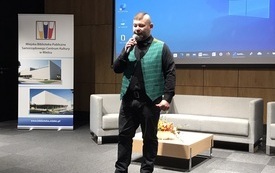 Mężczyzna w czarnych spodniach, koszuli i kamizelce w zieloną kratkę z mikrofonem w ręku.