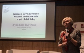 Kobieta w średnim wieku w okularach i kolorowej apaszce z mikrofonem w ręku. Na ekranie projekcyjnym  prezentacja multimedialna.