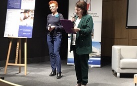 Dyrektor biblioteki wojewódzkiej stoi z mikrofonem i kartką w ręku. Obok kobieta w rudych włosach z mikrofonem. Na stojaku plakat z programem konferencji.