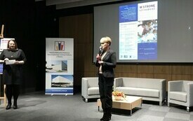 Kobieta w krótkich włosach z mikrofonem. W tle plakat z programem konferencji. Obok kobieta w czarnej sukience.