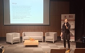 Kobieta w czarnej sukience w białe wzorki w okularach z mikrofonem w ręku. Na ekranie projekcyjnym prezentacja multimedialna.