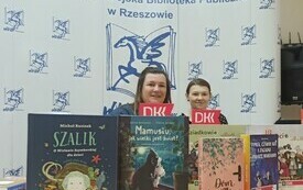 Okładki książek na stole, za książkami siedzą dwie kobiety z logo DKK. W tle logo pegaza Wojew&oacute;dzkiej i Miejskiej Biblioteki Publicznej w Rzeszowie.