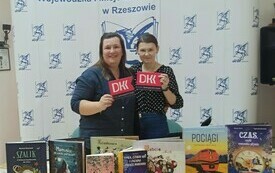 Dwie kobiety stoją obok siebie i tzrymają loga Dyskyusyjnych Klubów Książki. Na stole wystawka z książkami. W tle logo pegaza Wojewódzkiej i Miejskiej Biblioteki Publicznej w Rzeszowie.