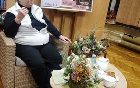 Kobieta siwowłosa w okularach siedząca na fotelu. Przed nią stoliki z kompozycjami kwiatowymi. 