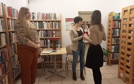 Regały w bibliotece, trzy kobiety stojące pośrodku. Pisarka przyjmuje prezent. 