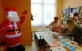Komisja konkursowa przegląda ułożone na stoliku prace konkursowe. Obok stolika postać Świętego Mikołaja.