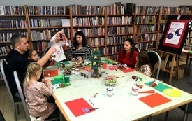 Dzieci wraz z opiekunami siedzą przy stole podczas i wykonują świąteczne dekoracje. W tle regału biblioteczne.