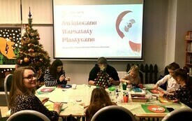 Przy stole grupa dzieci wraz z opiekunami wykonuje świąteczne dekoracje. W tle ekran projekcyjny, na kt&oacute;rym widnieje napis świąteczne warsztaty plastyczne.