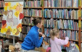 Bibliotekarka i dziewczynka zawieszają na sznurku obrazek. W tle regały z książkami. 