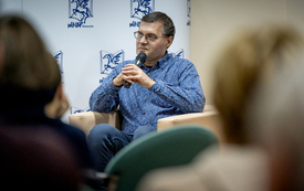 Mężczyzna w średnią wieku w koszuli w niebieskie wzory siedzi w fotelu. W ręku trzyma mikrofon.