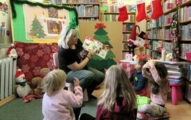 Kobieta z świąteczną opaską na głowie pokazuje siedzącym na dywanie dzieciom ilustracje w książce. Wok&oacute;ł regały z książkami i świąteczne dekoracje. 