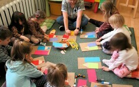 Dzieci wraz z prowadzącą spotkanie kobietą siedzą na dywanie. Uczestnicy spotkania wykonują prace plastyczne - papierowe kredki.