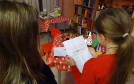 Dwie dziewczynki wpatrzone w tekst znajdujący się na kartce, kt&oacute;ra trzyma jedna z dziewczynek. Przed dziewczynkami stoliki i regał biblioteczny z książkami.