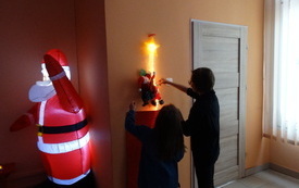 Kobieta i dziewczynka stoją przed ścianą, na kt&oacute;rej znajduje się ozdoba: Mikołaj wspinający się po łańcuchu do gwiazdki. Obok dmuchana postać Mikołaja. 