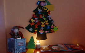 Na ścianie choinka ozdobiona papierowymi o filcowymi ozdobami. Poniżej zaświecona lampka, figurka Mikołaja i świąteczne książki. 