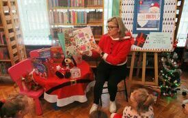 Kobieta w mikołajkowym stroju pokazuje zgormadzonym w bibliotece dzieciom książkę o świętach. 