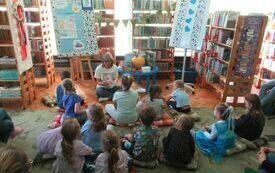 Na dywanie w bibliotece siedzi grupa dzieci. Przed nimi kobieta w okularach z książką w ręku. Wok&oacute;ł regały z książkami.