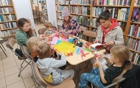 Przy stoliku grupa dzieci wraz z opiekunami wykonuje świąteczne dekoracje z papieru. W tle regały biblioteczne.