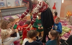 Młoda kobieta w świątecznym swetrze ze świąteczną skarpetą wśr&oacute;d siedzących na dywanie dzieci.