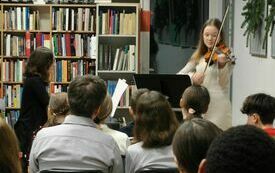 Młoda dziewczyna z długimi włosami gra na skrzypcach. Przed nią zgromadzona publiczność.